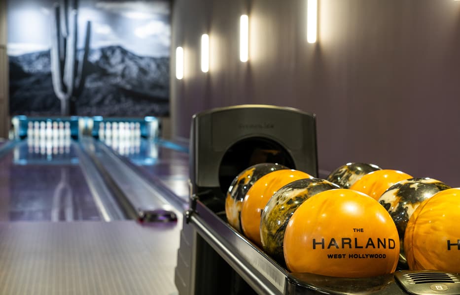 Close-up of a set of bright orange and black bowling balls on a rack with The Harland West Hollywood branding in front of two polished bowling lanes inside a luxury amenity space.