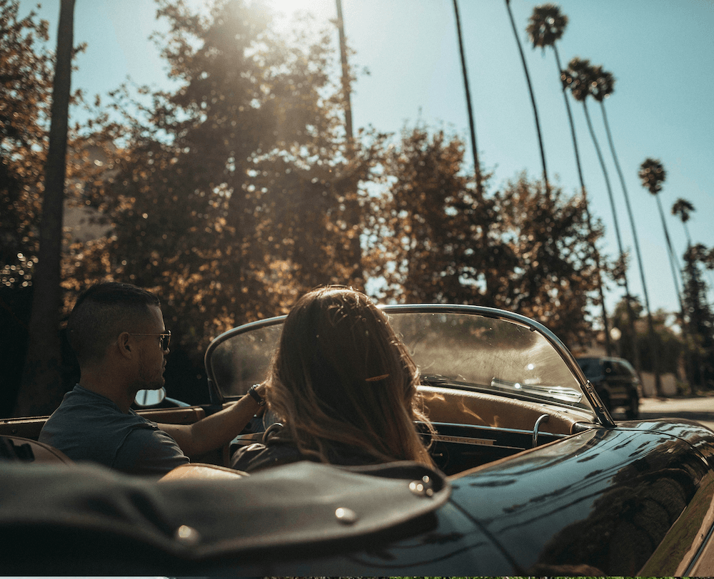 Two people riding in a convertible car down a sunny street lined with tall palm trees and leafy trees, with sunlight filtering through the foliage.
