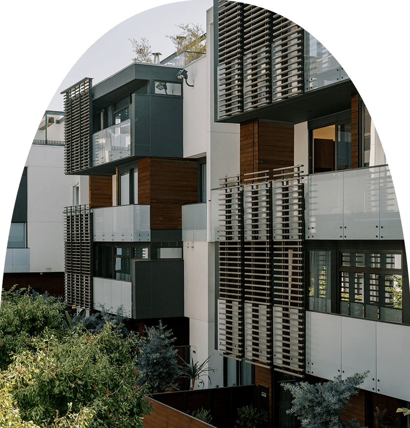 Exterior detail of a modern residential building façade showing staggered balconies with glass railings and wooden slat screens, with greenery and landscaped shrubs in the foreground.