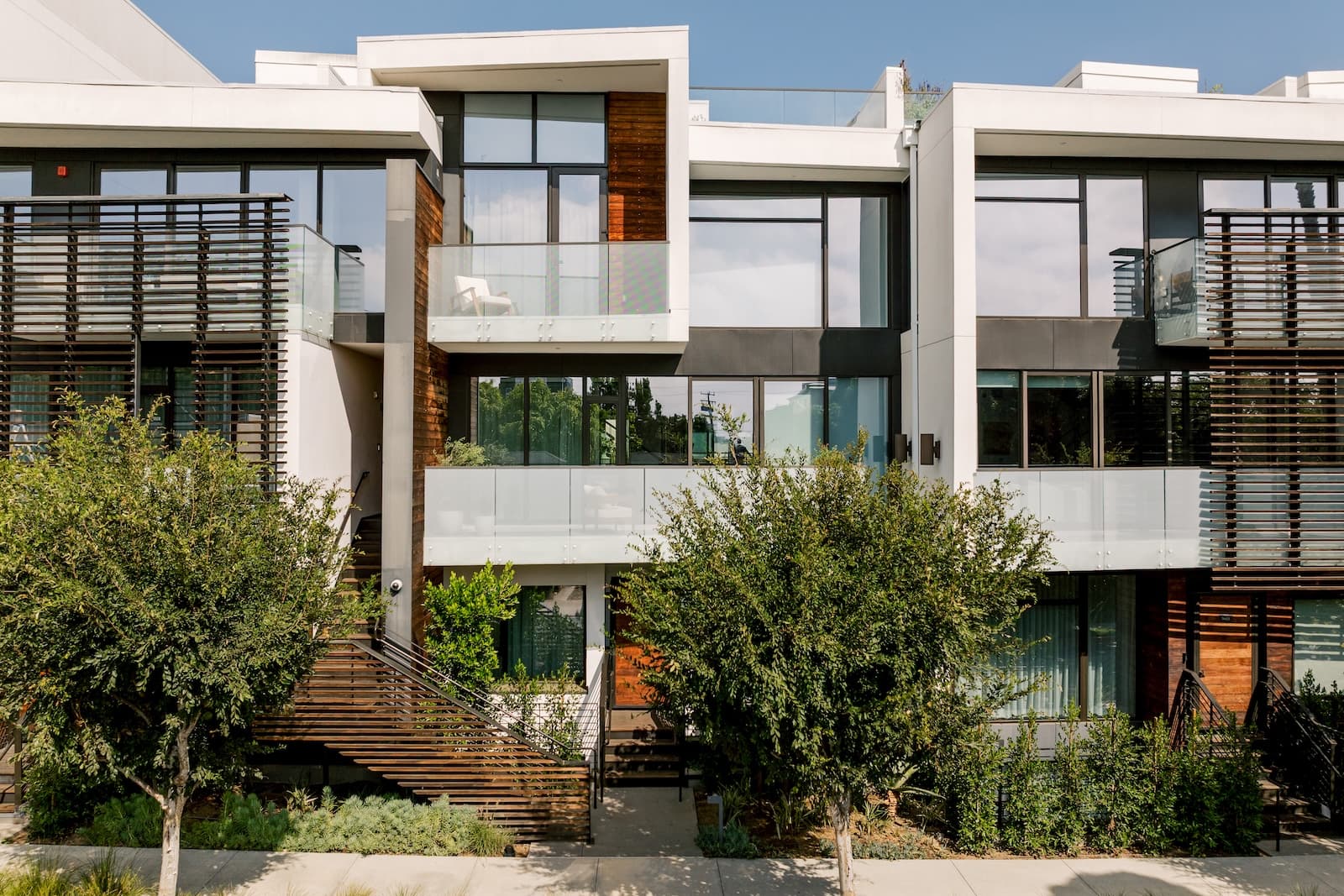 Exterior of a modern multi-story residential building with large glass windows and balconies, featuring horizontal wood panel accents on the façade and leafy trees in front