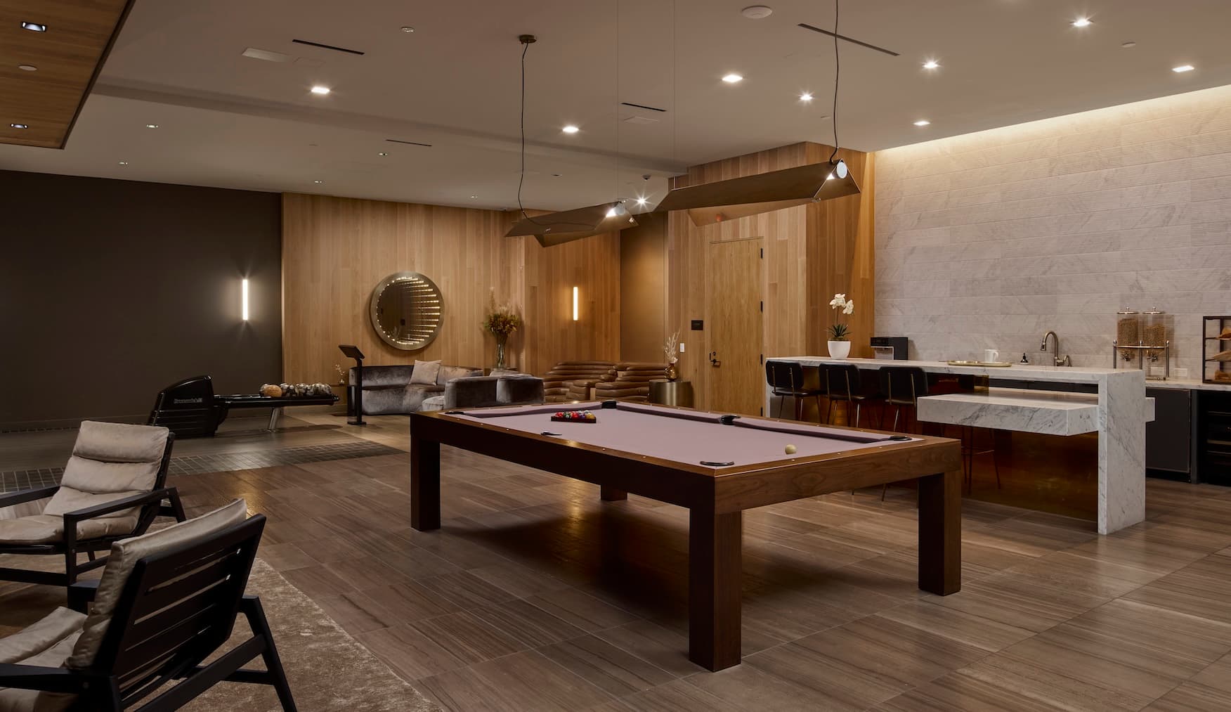 Modern luxury amenity lounge with warm wood-paneled walls, a central pool table, lounge seating, and a marble-topped bar area with high stools. Contemporary pendant and recessed lighting illuminate the open space, which also features a round wall mirror, decorative plants, and a fitness area with exercise equipment in the background. The design combines rich wood tones, stone finishes, and soft furnishings to create an upscale, inviting atmosphere.