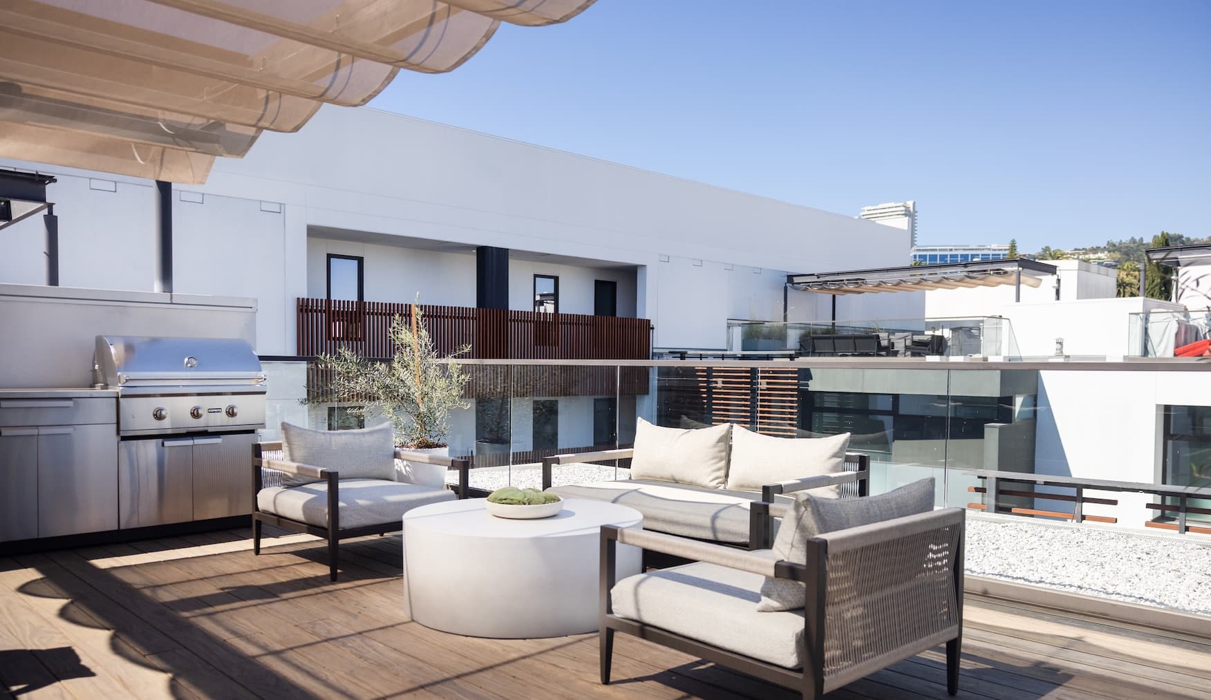 Contemporary rooftop terrace with outdoor lounge seating, a round coffee table, and a built-in stainless steel gas grill. The space features wood-look decking, glass railings, and retractable fabric shade canopies, overlooking a modern apartment courtyard with white façades and wood-accent privacy screens under a clear blue sky.