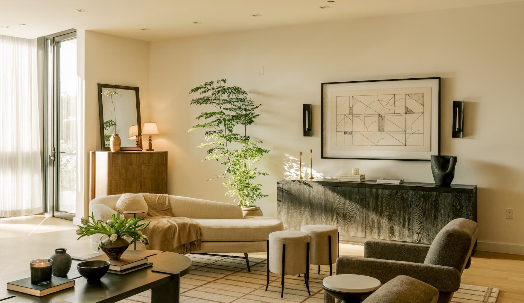 Warm, sunlit modern living room with neutral furnishings, a cream chaise sofa, dark wood console, sculptural armchair, large leafy indoor tree, abstract framed artwork, and layered coffee tables on a geometric rug.