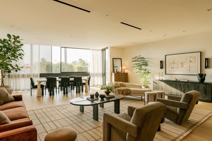 Spacious, light-filled living and dining area in a luxury residence at The Harland West Hollywood with a large sofa and armchairs around a central coffee table, a dining table with chairs by floor-to-ceiling windows, potted plants, and contemporary décor in neutral tones.