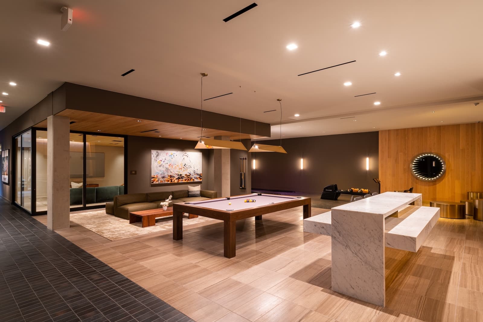 Residents lounge with a pool table in the center, a large sectional sofa and coffee table to the left, modern lighting overhead, and a long marble-topped table on the right in a warm, stylish social space.