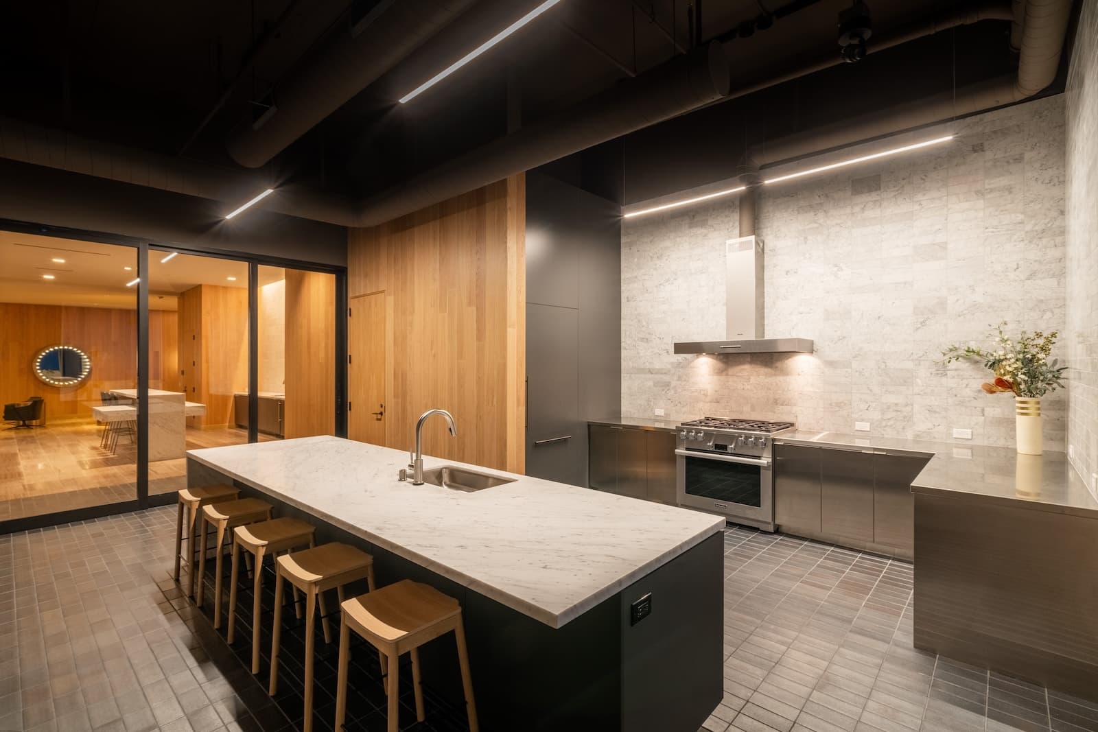 Demonstration kitchen amenity with a long marble island countertop, bar stools, stainless steel appliances, and wood-accent cabinetry in a modern social space.