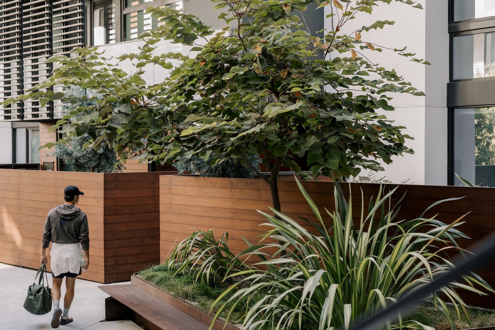 Man walking through a landscaped outdoor courtyard at The Harland West Hollywood, with green plants, trees, and modern building façades with wood paneling and windows in the background.