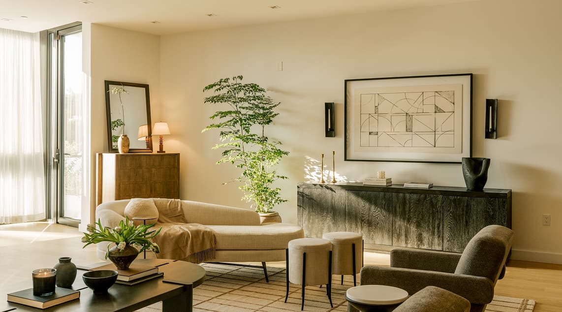 Sunlit modern living room with curved sofa, neutral furnishings, indoor greenery, abstract wall art, and floor-to-ceiling windows creating a warm, minimalist interior.