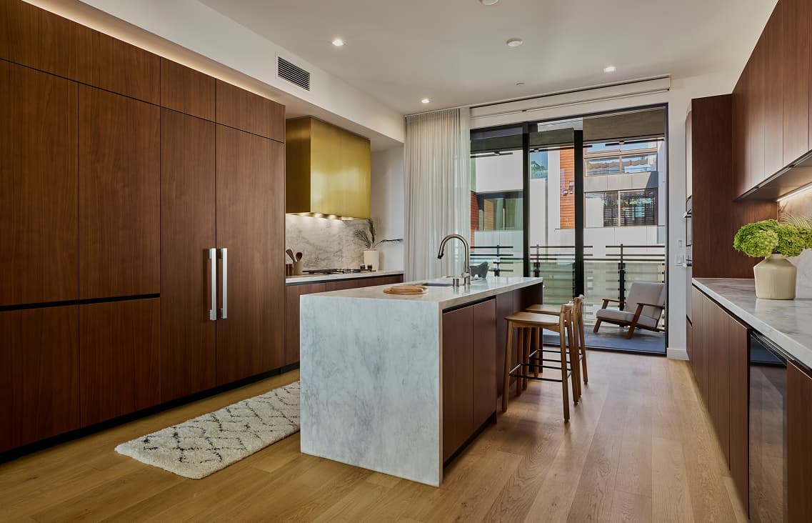 Open-plan kitchen and entrance to outdoor terrace with a central island topped in white stone and wood base, a long wood cabinet wall with a gold range hood above the stove, terraced windows let in natural light