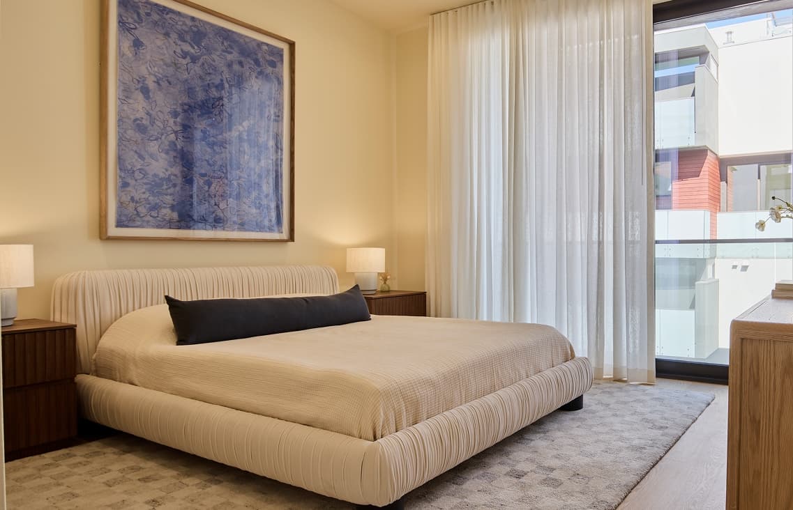 A modern primary bedroom featuring a bed and minimal furnishings, with large sliding glass doors that open to a private balcony. The space emphasizes indoor-outdoor living, allowing natural light to enter and providing direct access to an outdoor seating area