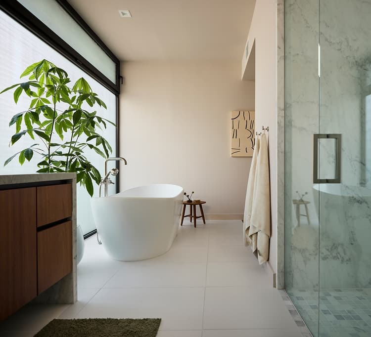 Primary bathroom with a freestanding white bathtub in the center, a marble-topped vanity with sink and large mirror on one side, a glass-enclosed shower on the other, and light coming through a frosted window.