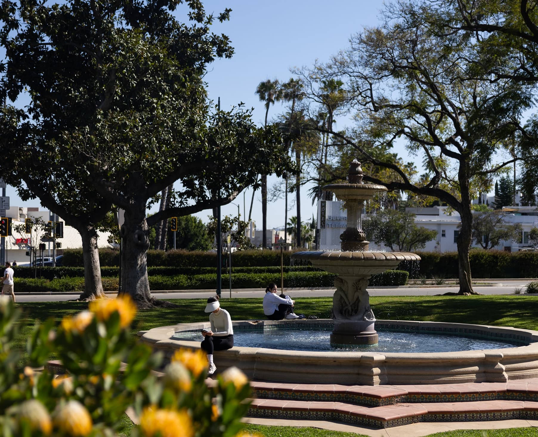 Tree-lined Beverly Hills park with walking paths, a fountain, green lawns, and landscaped gardens.