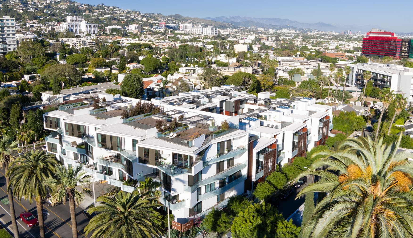 Aerial view of The Harland West Hollywood condominium building with modern white facades, glass balconies, and rooftop terraces, set among city streets, greenery, and surrounding buildings under a clear sky.