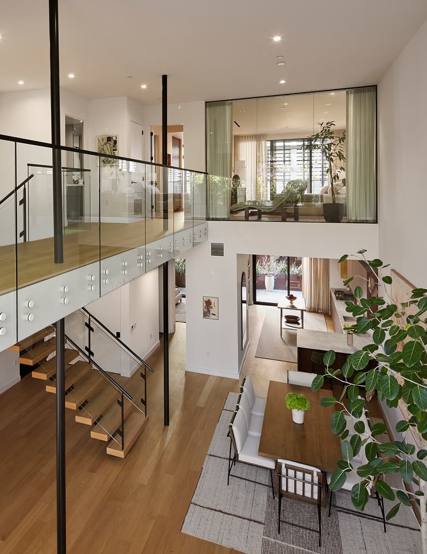A wide-angle interior view of a modern residence at The Harland, showing an open-plan layout that combines the living room and kitchen. The space features high ceilings, large floor-to-ceiling windows allowing natural light, and contemporary finishes. Seating and kitchen areas are arranged in a clean, cohesive design that emphasizes openness and flow.