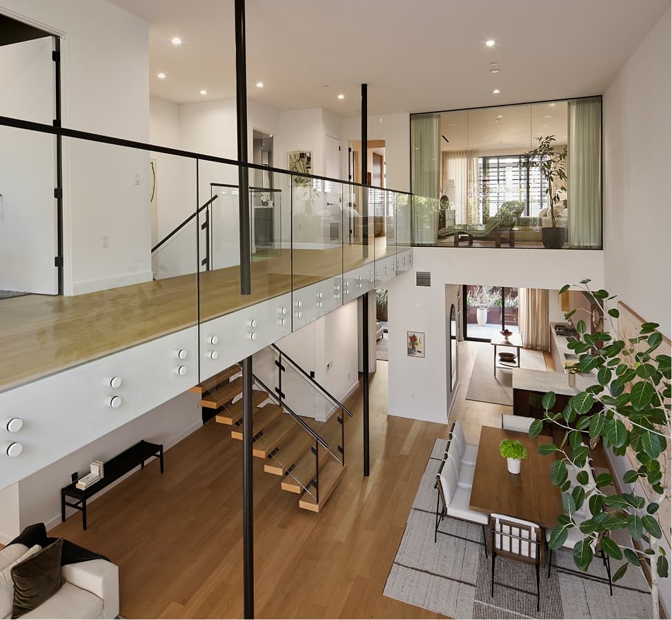 A wide-angle interior view of a modern residence at The Harland, showing an open-plan layout that combines the living room and kitchen. The space features high ceilings, large floor-to-ceiling windows allowing natural light, and contemporary finishes. Seating and kitchen areas are arranged in a clean, cohesive design that emphasizes openness and flow.