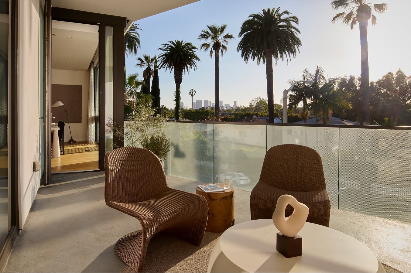 Sunlit balcony of a modern West Hollywood residence with a glass railing, outdoor seating including a cushioned sofa and woven chairs around a round coffee table, potted plants, and tall palm trees and city buildings visible in the background.