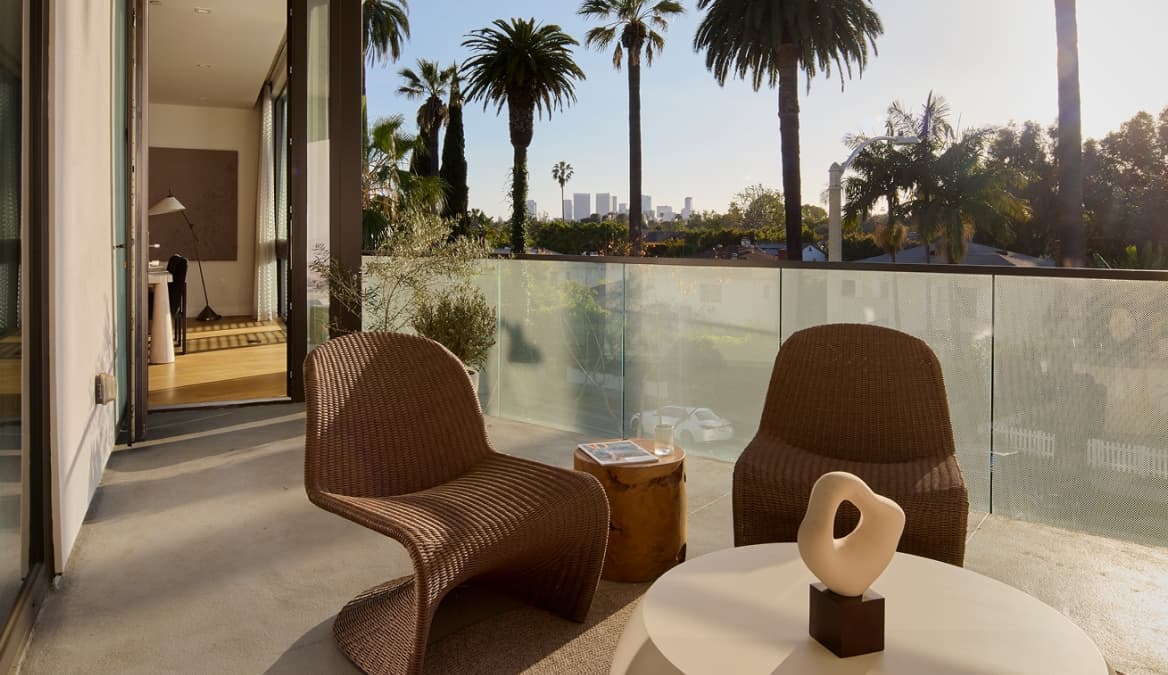 Sunlit balcony of a modern West Hollywood residence with a glass railing, outdoor seating including a cushioned sofa and woven chairs around a round coffee table, potted plants, and tall palm trees and city buildings visible in the background.