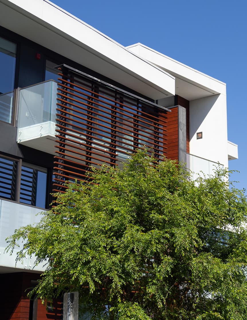 An exterior view of The Harland showing a contemporary residential building integrated with landscaped surroundings. The structure features multiple levels with balconies, layered façade elements, and greenery that softens the architecture. The design emphasizes indoor-outdoor living and a connection to the surrounding environment.