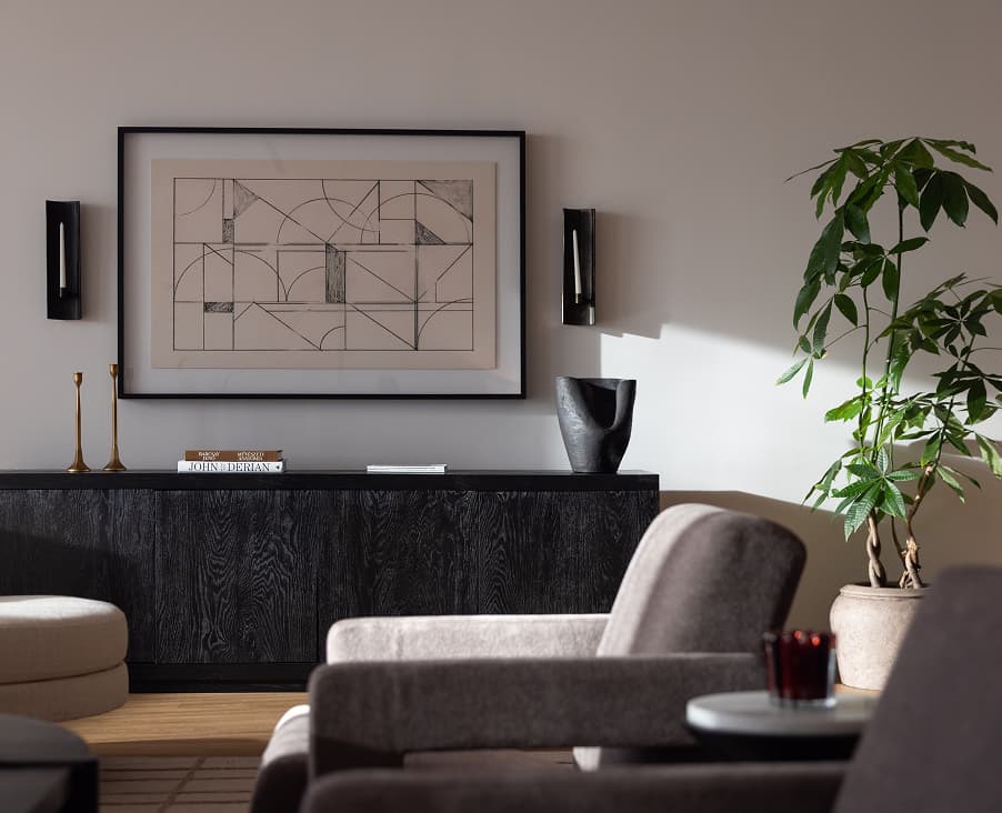 Interior sitting area of a primary bedroom in a luxury residence showing a wooden sideboard with artwork above it, decorative objects on top, a leafy potted plant to the left, and curved cushioned seating in the foreground.