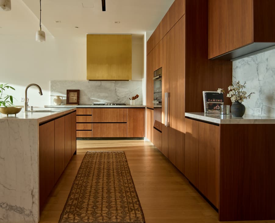 Modern kitchen interior with wood cabinetry and a stove with a gold range hood covering the back wall, a large island with a white countertop and sink in the foreground, and light wood flooring.