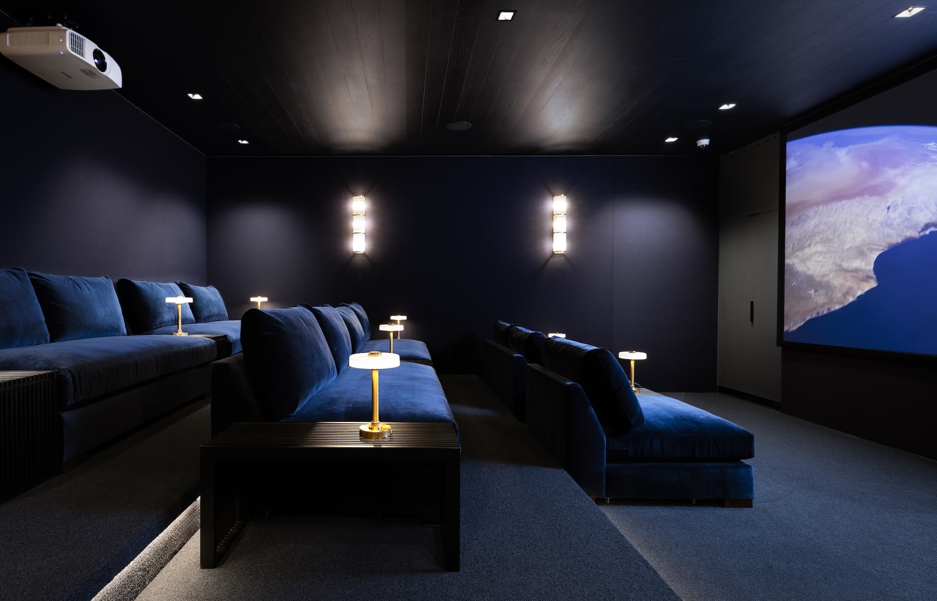 Luxurious private screening room with tiered rows of deep blue velvet sofas facing a large projection screen. Dark navy walls and ceiling create a cinematic atmosphere, accented by warm brass table lamps and wall sconces. A ceiling-mounted projector and minimalist design give the space an elegant, high-end home theater feel.